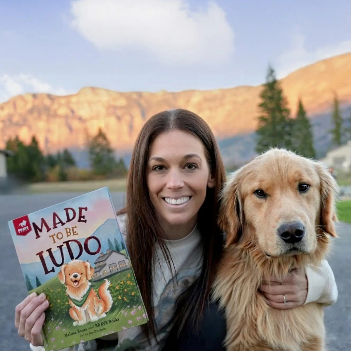 Melissa Souza holding a book titled 'Made to be Ludo' with a dog in a mountainous landscape.