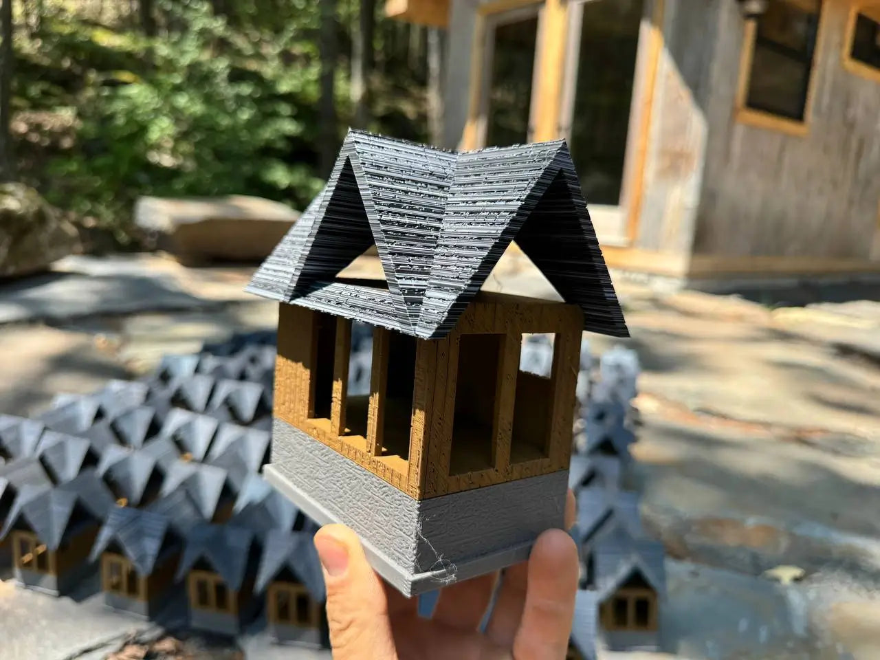 Hand holding a small 3D-printed model of a building with a background of more models and a real building.