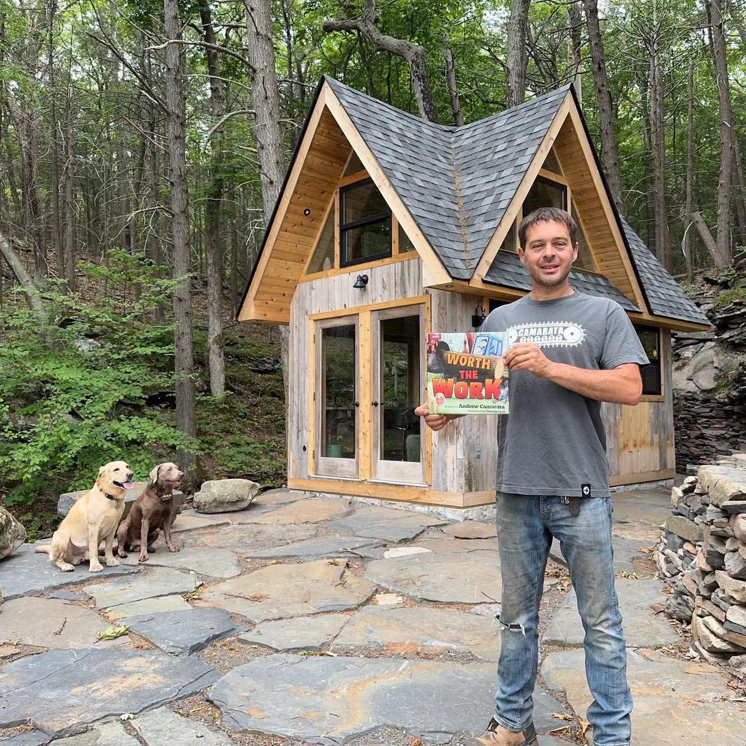 Andrew Camarata with his book in front of a small wooden cabin in the woods with two dogs.