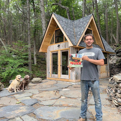 Andrew Camarata with his book in front of a small wooden cabin in the woods with two dogs.