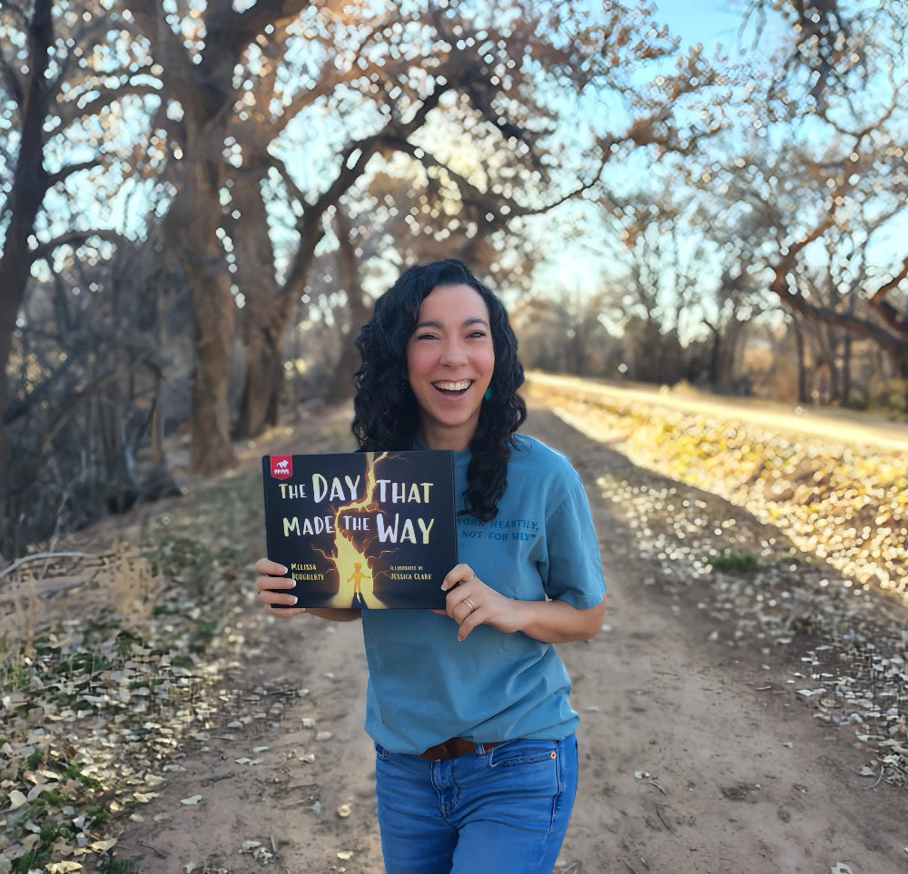 Melissa Dougherty holding a book titled 'The Day That Made the Way' in a scenic outdoor setting.