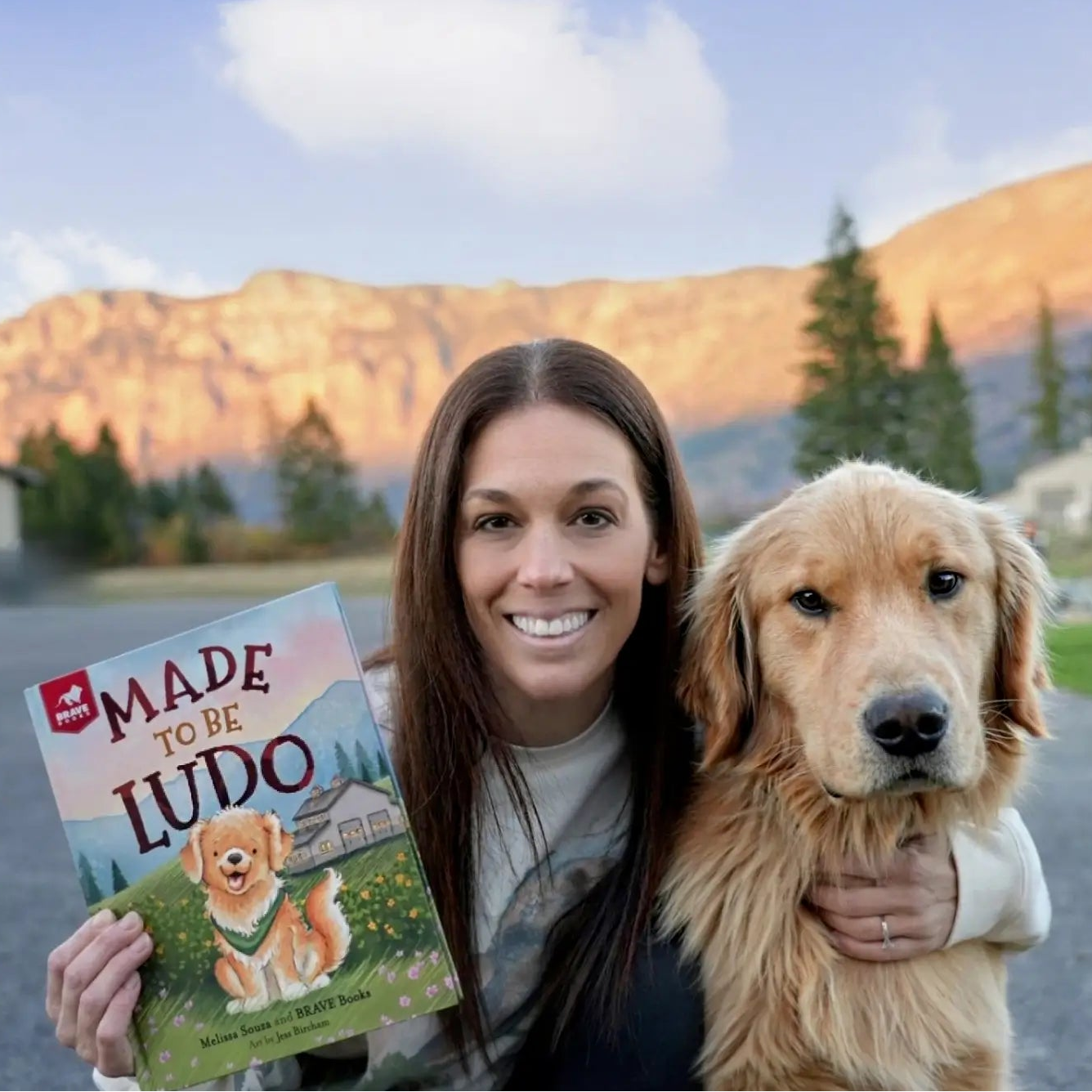 Melissa Souza holding a book titled 'Made to be Ludo' with a dog in a mountainous landscape.